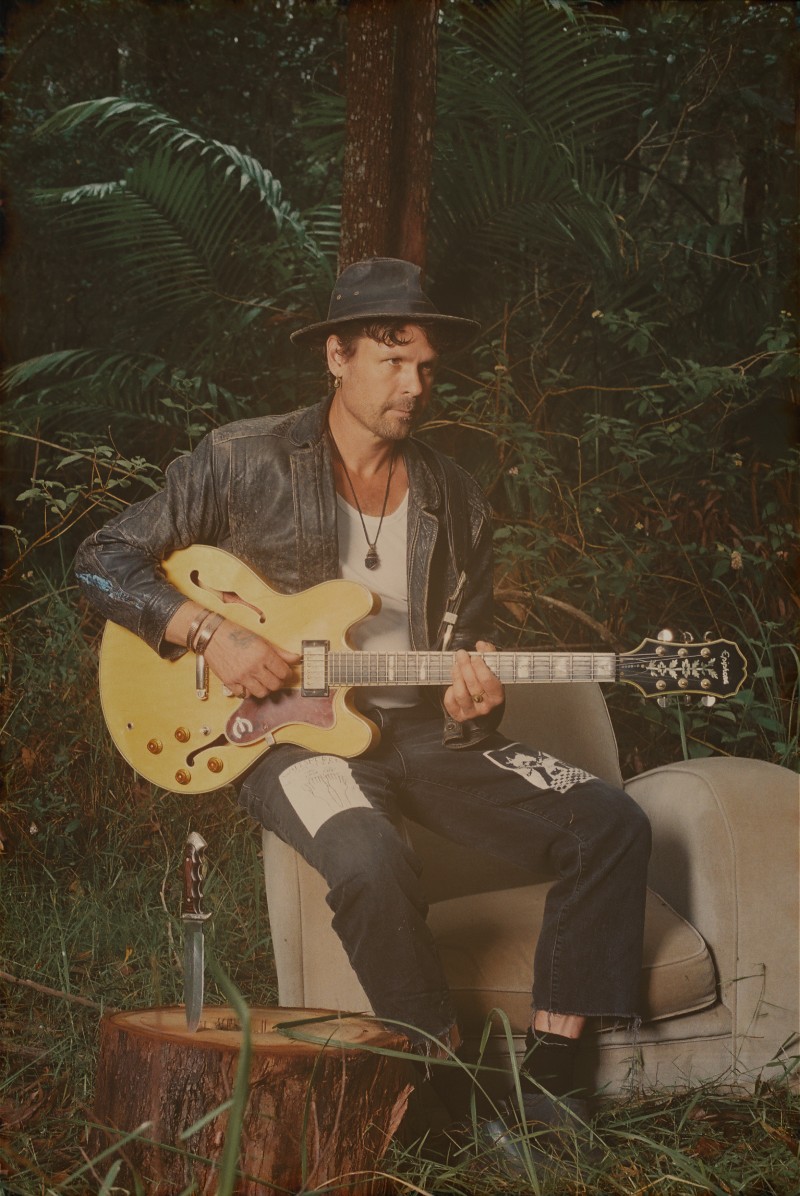 WaltSin on an armchair in subtropical bush, guitar in hand, with a knife on the stump beside him — warm amber tones