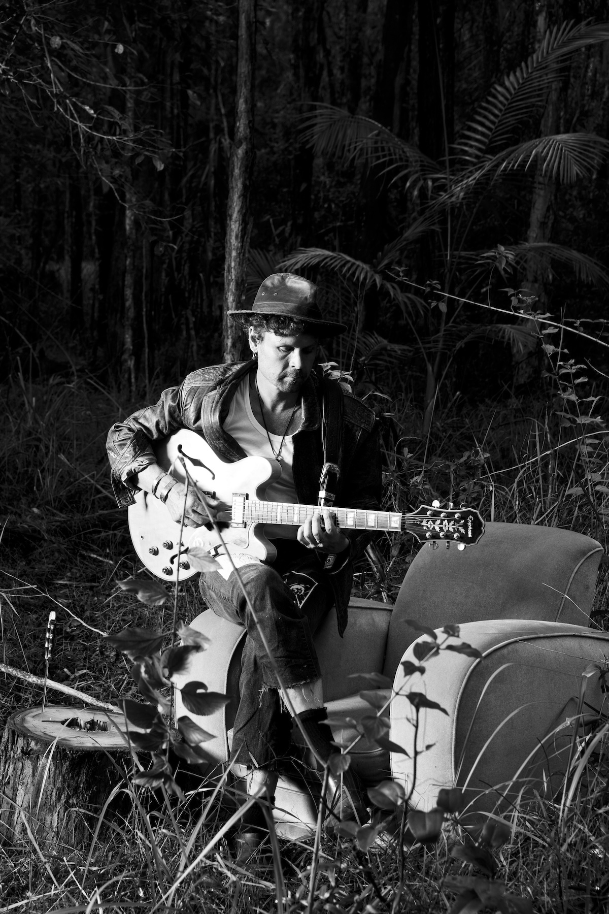 WaltSin in the field — black and white portrait, seated in armchair in dark subtropical bush, playing white guitar
